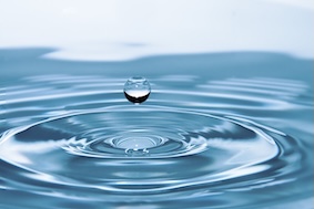 A single water droplet suspended above a calm surface, with concentric ripples spreading below it.