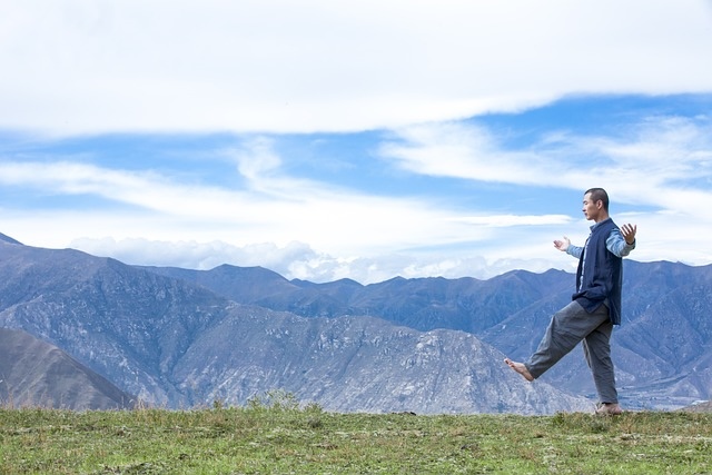 A man practicing walking meditation on a grassy field, with mountains and a partly cloudy sky in the background. The pose emphasizes balance, breath, and mindful movement in harmony with nature.